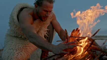 Man in animal hide blowing at burning fire then warming hands in evening in barbarian camp - slow motion