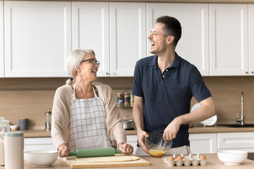 Cheerful son helping senior mom to make homemade pie, cookies. Mature mother and adult child man...
