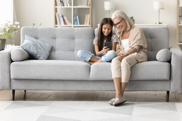 Happy grandmother and preteen granddaughter using Internet app on cellphone, sitting on couch, talking on video call together, enjoying online communication at cozy home