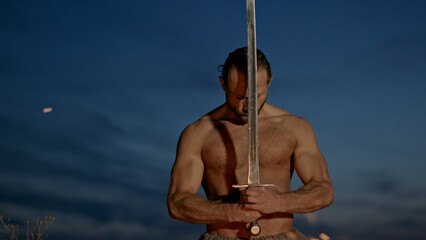 Barbarian warrior with sword near campfire. Muscular shirtless man with sword kneeling near burning fire against cloudy evening sky in nature