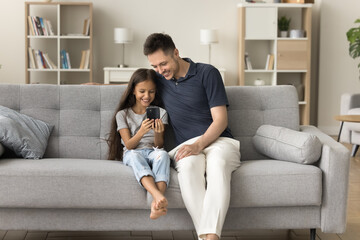 Happy positive father and little kid girl using smartphone for online communication, playing on digital device together, relaxing on comfortable home couch, enjoying leisure, Internet technology