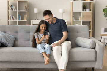 Obraz premium Happy positive father and little kid girl using smartphone for online communication, playing on digital device together, relaxing on comfortable home couch, enjoying leisure, Internet technology