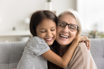 Excited loving granddaughter kid and happy caring grandmother hugging on home couch with love,...