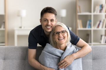 Handsome loving adult son man hugging happy mature mom from behind, wrapping mothers shoulder in...