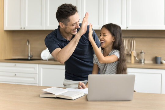Excited schoolkid girl and happy dad giving high five over learning textbook and laptop on kitchen table, laughing, celebrating education success, achievement, enjoying studying, teaching
