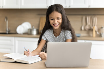 Happy preteen schoolchild girl studying online at home, doing school homework task, sitting at table and learning book, writing notes, looking at display, watching lesson on Internet, smiling