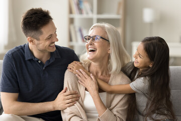Happy adult son and little granddaughter girl hugging happy grandma with love, affection, smiling, laughing, celebrating family event, holiday, mothers day, spending funny leisure time at cozy home