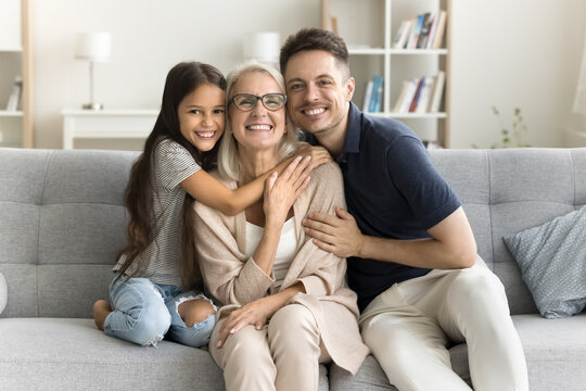Happy sweet granddaughter kid and young dad hugging beloved grandma. Cheerful preteen girl, father and grandmother posing for family picture on home couch, looking at camera, smiling