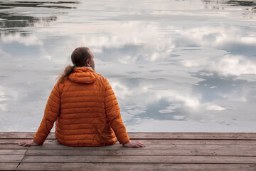 Lonely man in orange hiking down jacket sitting on the wooden pier