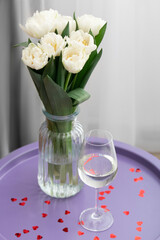 A bouquet of white tulips in a vase and a glass of champagne on a purple table decorated with red hearts. Valentine's Day, birthday, March 8. Top view.