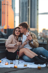 Fototapeta premium Surprise date on rooftop with urban cityscape and skyscrapers on background. Happy young loving couple drinking wine having romantic candlelit dinner celebrating anniversary or Valentines Day