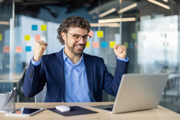 Mature experienced businessman in business suit is looking at successful achievement results, boss with laptop inside office holding hands up, gesture of success, triumph, online notification laptop.