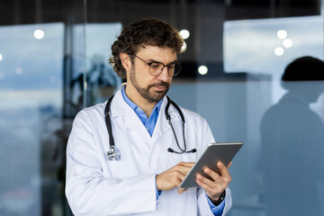 Mature successful doctor inside medical office, clinic office, man in white medical coat and stethoscope uses tablet computer to consult patients remotely online.