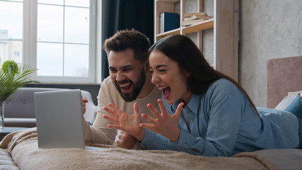 Excited Caucasian married couple lying on bed at home win laptop achievement read amazing news...