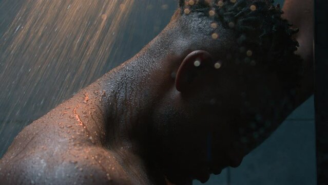 Close up sad depressed African American naked man under falling hot water in shower post workout stress impotence mental problems depression thinking of mistake frustrated guy washing in dark bathroom