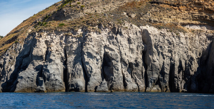 Weathered seaside rock face texture with parts of green and blue water. Aged volcanic stone wall surface background pattern with cracks and scratches. Banner. Ischia Island, Italy.