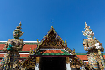 Fototapeta na wymiar Colorful entrance to building in the Grand Palace, Bangkok, Thailand. Intricate gold decorations. Pair of Giant statues guarding the door. Blue sky behind. 
