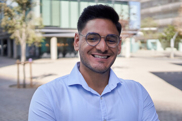 Obraz premium Portrait young adult Latin business man or commercial outside office center looking smiling at camera. Male freelance worker in shirt poses cheerful for job photo outdoors. Copy space staff people
