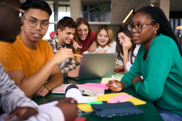 Selective focus on a smiling male young university student using laptop in the cafeteria on campus with his female classmates. Academic multiracial people studying together on the faculty building