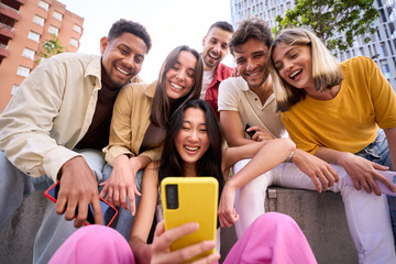 Young group of gen z people having fun using cell phone together outside watching something funny at mobile app. Cheerful community of student enjoying social media content. Multiethnic friends - obrazy, fototapety, plakaty