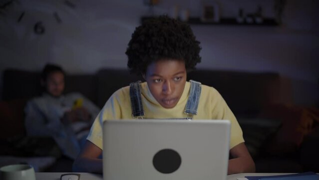 Young Freelance Afro Woman Concentrating Using Laptop At Night Room . African American Female Staring At Computer Screen. Millennial Black Person Working Online From Home. Unfocused Man In Background