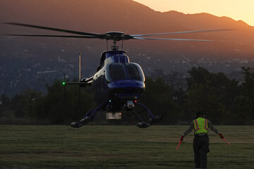 An aircraft marshaler guides an unmarked helicopter while landing in a field during the early morning, with mountains in the background.