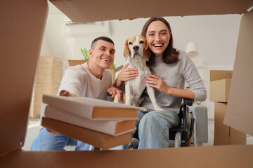 Young man and his wife in wheelchair with Beagle dog putting books into box on moving day, view...
