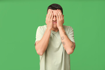 Handsome ashamed young man covering face with hands on green background