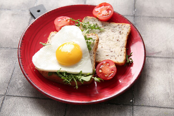 Plate with tasty fried egg, toasts, tomatoes and arugula on grey tile background, closeup
