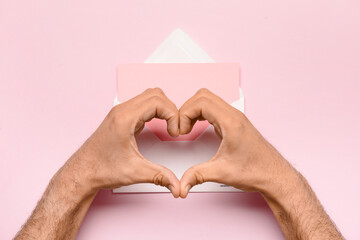 Male hands making heart shape with envelope and blank card on pink background. Valentine's Day...
