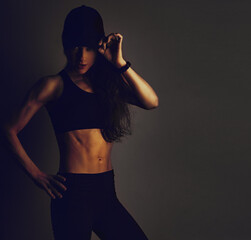 Sport muscular woman posing in black sport bra and cap showing the shoulders, blades and arms, standing on dark shadow studio background. Back body view and profile face. Sporty healthy toned vintage