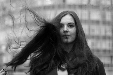 Black and white poster. Street photo. Beautiful young white childfree brunette woman. Glass wall of building with windows. Wind in loose hair, covering face. Concept of freedom, beauty, naturalness