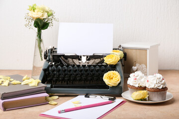 Vintage typewriter with books, cupcakes and flowers on beige grunge table near white wall