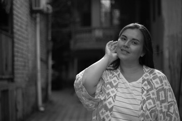 Fototapeta premium Black and white poster. Street photo. One tanned young childfree white brunette woman in pink shirt looks at camera and strokes her hair on sunny day in courtyard of southern house. Travel, tourism