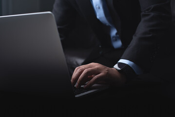 Business man in black suit working on laptop computer, hand typing on keyboard on table at office with dark background, online working, close up