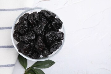 Sweet dried prunes in bowl and green leaves on white textured table, top view. Space for text