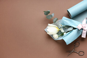 Mini bouquet of beautiful spring flowers in wrapping paper with scissors on brown background. International Women's Day