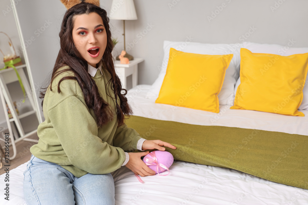 Sticker Surprised young woman in bunny ears with Easter egg under blanket on bed at home