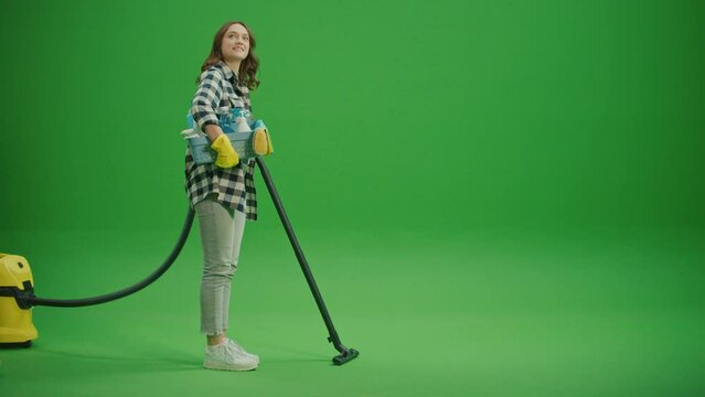 Green Screen. A Smiling Young Woman Housewife Walks with a Vacuum Cleaner and a Box with Cleaning Products.The Housewife is Vacuuming the Floor.Minimalist Cleaning with Multi-functional Appliances.