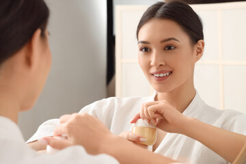 Young Asian woman with jar of facial cream near mirror in bathroom, closeup