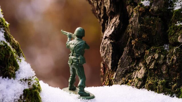 A toy soldier stands on a brown, green mossy, snowy tree branch in the frozen, cold, frosty winter weather. In the background, the nature is blurred.
