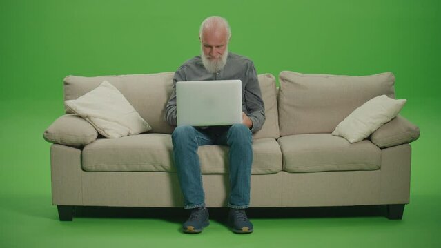 Green Screen. A Smiling Old Man Sitting on a Sofa and Works on a Laptop. A Smart Elderly Man, Freelancer, Browsing on the Internet Using a Laptop. Security and Privacy Concerns for Senior Tech Users.