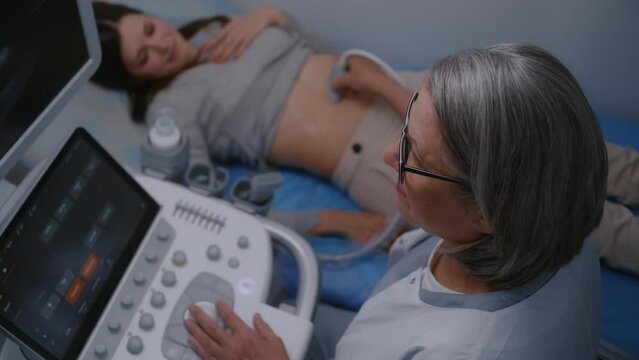 Female medical specialist, doctor performs check up of abdominal organs to female patient using ultrasound machine with digital monitor. Adult woman undergoes sonography diagnostics in modern clinic.