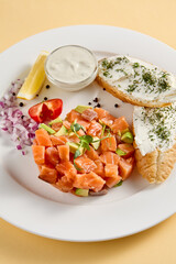 Salmon and avocado tartare with crispy bread and cream cheese, elegantly plated with lemon and dill on a white plate against a yellow background