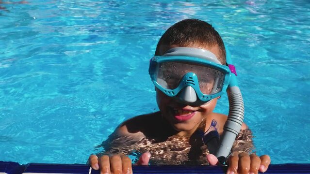 A child wearing swimming goggles with a snorkel swims up to the edge of a blue water pool. Summer vacation on the water. Vacations and children's rehabilitation.