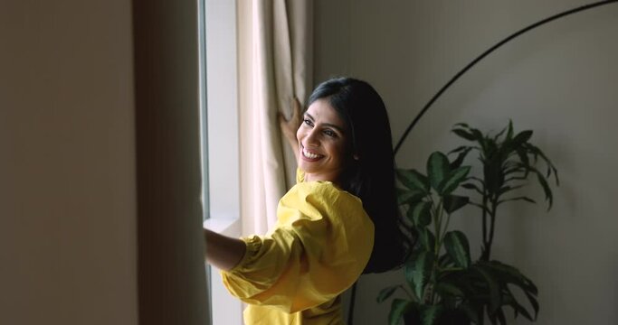 Side view beautiful Indian woman opening curtains, admiring city view from own or rented apartment, enjoy holidays, standing near window, welcoming new day. Good morning, bank mortgage, own flat owner