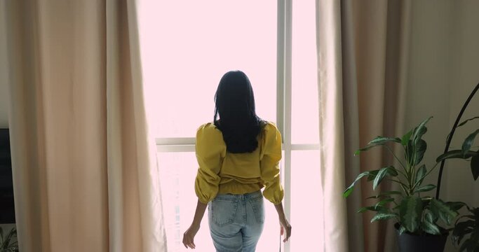 Rear back view happy young woman opening curtain standing alone in luxury apartment or modern hotel looking through panoramic window enjoying city view in the morning. New dwelling, ownership, travel