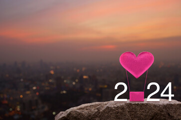 Pink fabric heart love air balloon with 2024 white text on rock mountain over cityscape on warm...