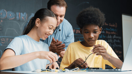 Caucasian teacher talking about electric tool while student fixing robotic model on table with...