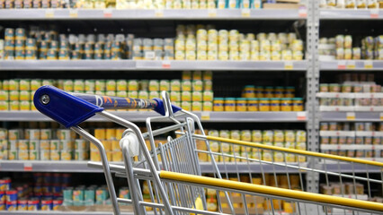 Close-up of a shopping trolley in a grocery store against a blurred background of many tins of...